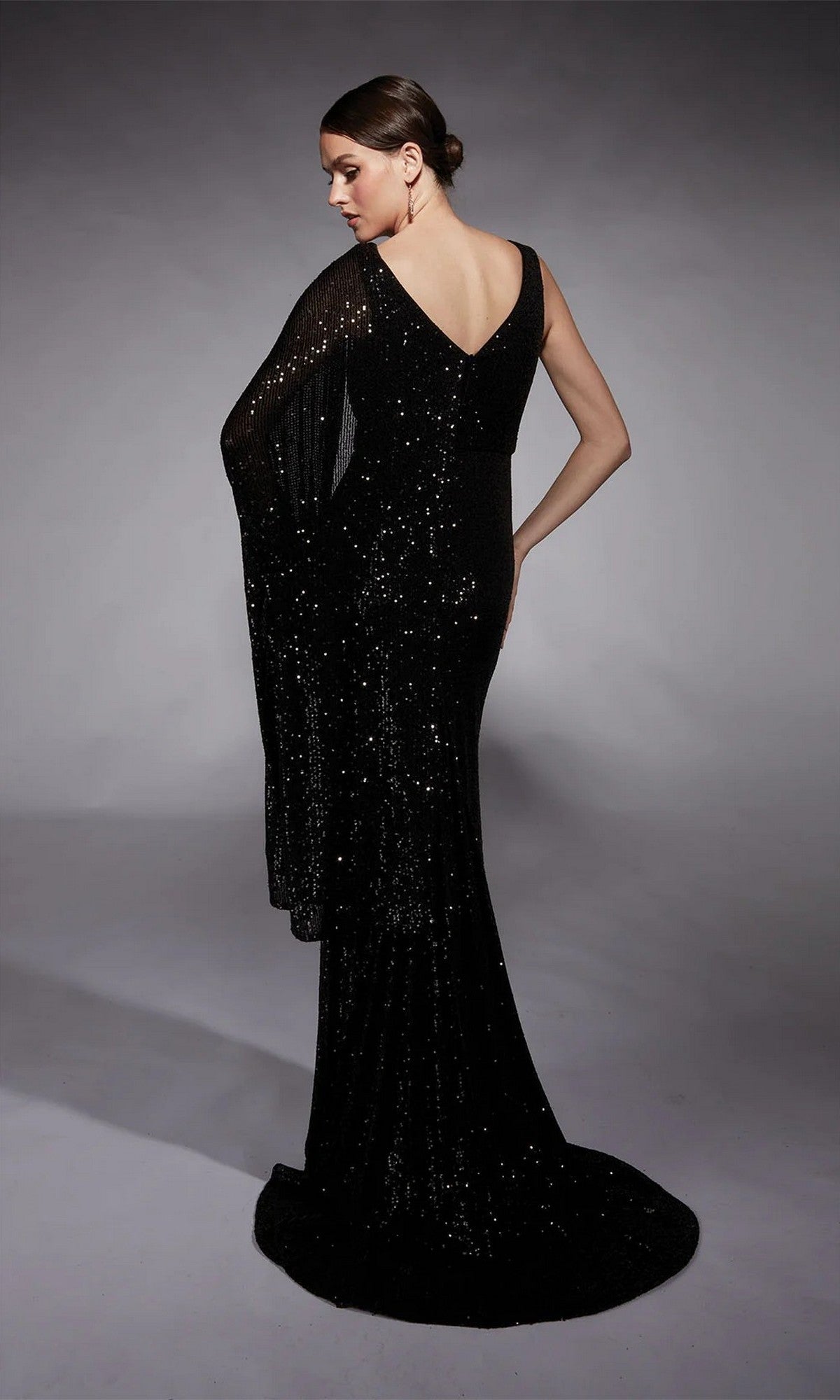 A woman stands with her back to the camera against a neutral background, wearing the Alyce 27718 long formal dress—a floor-length sequin gown with a one-shoulder cape sleeve.